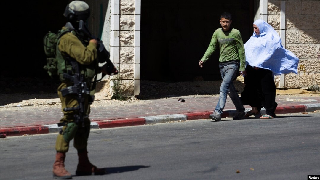 Palestinians walk near an Israeli soldier as he stands guard during an operation to locate three Israeli teens in the West Bank City of Hebron, June 17, 2014.
