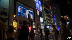 Japanese Prime Minister Yoshihide Suga, on a screen, speaks at a news conference in a live broadcasting shown in a big screen on a building as people walk across an intersection in the Shinjuku district of Tokyo on Thursday, March 18, 2021. Japan…