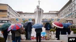 Pessoas rezam debaixo de chuva junto à estátua de João Paulo II no Hospital Universitário Gemelli, onde o Papa Francisco está hospitalizado com pneumonia, em Roma, a 1 de março de 2025.