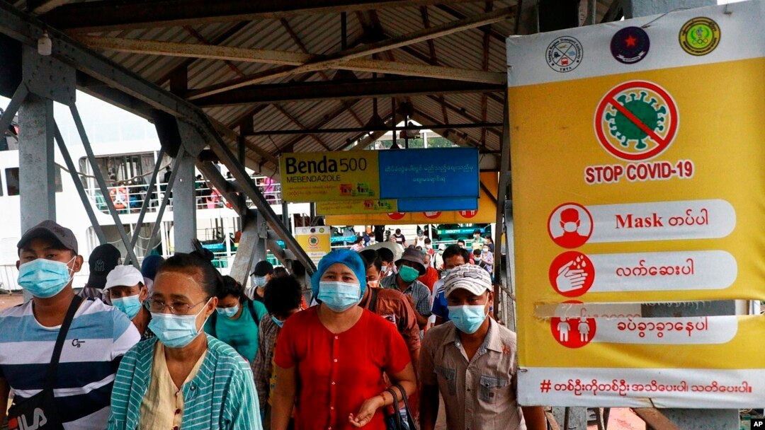Penumpang feri mengenakan masker di tengah pandemi COVID-19, berjalan melewati poster "Hentikan COVID-19" di dermaga Pansodan, Yangon, Myanmar. (AP)