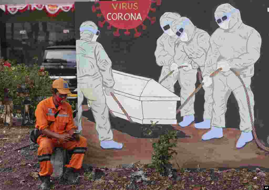 A street sweeper checks his mobile phone as he takes a break near a coronavirus-themed mural in Jakarta, Indonesia.